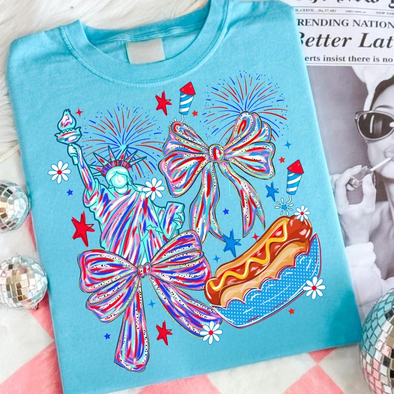 May include: Light blue t-shirt featuring a patriotic design with the Statue of Liberty, fireworks, bows, a hot dog, and stars. The graphic is in red, white, and blue colors, perfect for Independence Day celebrations.