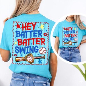 K&ouml;nnte beinhalten: Hellblaues T-Shirt mit Baseball-Motiv. Der Aufdruck zeigt die Worte "HEY BATTER BATTER SWING" in Rot und Blau, zusammen mit einem Baseball, Schl&auml;ger und Handschuh. Das Design ist von einem wellenf&ouml;rmigen Rand umgeben.