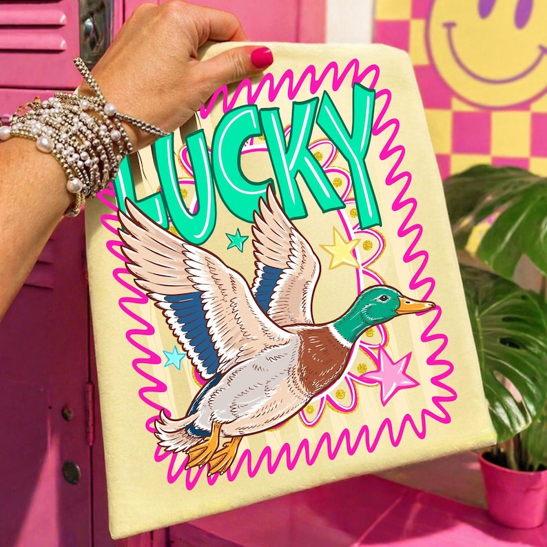 May include: A light yellow rectangular graphic featuring a flying duck illustration with the word "LUCKY" in green text. The duck has brown, blue, and white feathers. Pink stars and a pink wavy border surround the design.