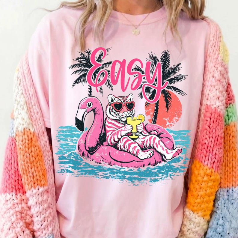 K&ouml;nnte beinhalten: Hellrosa T-Shirt mit einem Tiger mit Sonnenbrille auf einem rosa Flamingo-Schwimmer, dem Wort "Easy" dar&uuml;ber. Das Design zeigt Palmen und einen Sonnenuntergang. Es vermittelt eine entspannte Sommerstimmung.