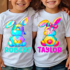 Pode incluir: Duas camisetas brancas com desenhos de coelhos tie-dye. O coelho à esquerda está usando um boné de beisebol ao contrário e o coelho à direita tem um laço rosa. O texto "ROBERT" está na camiseta da esquerda e "TAYLOR" está na camiseta da direita.