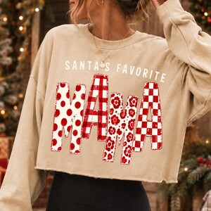 K&ouml;nnte beinhalten: Ein beiges, lang&auml;rmeliges, bauchfreies Sweatshirt mit dem Schriftzug SANTA'S FAVORITE MAMA in Wei&szlig; und Rot. Das Wort MAMA ist in gro&szlig;en Blockbuchstaben gedruckt, wobei jeder Buchstabe ein anderes rot-wei&szlig;es Muster wie Punkte, Karos und Blumen aufweist.