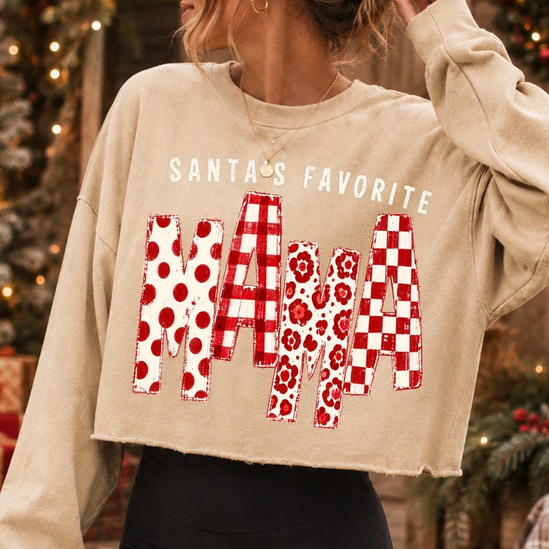 K&ouml;nnte beinhalten: Ein beiges, lang&auml;rmeliges, bauchfreies Sweatshirt mit dem Schriftzug SANTA'S FAVORITE MAMA in Wei&szlig; und Rot. Das Wort MAMA ist in gro&szlig;en Blockbuchstaben gedruckt, wobei jeder Buchstabe ein anderes rot-wei&szlig;es Muster wie Punkte, Karos und Blumen aufweist.