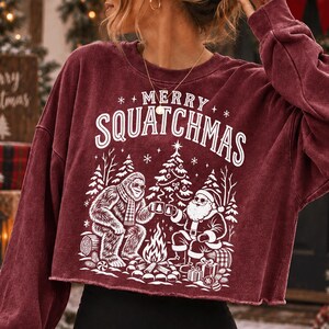 K&ouml;nnte beinhalten: Ein bordeauxrotes Langarm-Sweatshirt mit Rundhalsausschnitt und offenem Saum. Auf der Vorderseite befindet sich ein wei&szlig;er Aufdruck von Bigfoot und dem Weihnachtsmann am Lagerfeuer, dar&uuml;ber der Text MERRY SQUATCHMAS in einer geschwungenen Schrift.