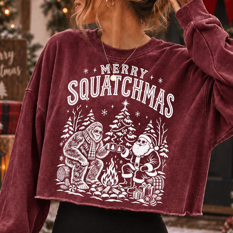 K&ouml;nnte beinhalten: Ein bordeauxrotes Langarm-Sweatshirt mit Rundhalsausschnitt und offenem Saum. Auf der Vorderseite befindet sich ein wei&szlig;er Aufdruck von Bigfoot und dem Weihnachtsmann am Lagerfeuer, dar&uuml;ber der Text MERRY SQUATCHMAS in einer geschwungenen Schrift.