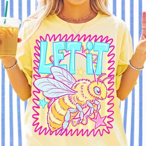 May include: Yellow t-shirt with the text "LET IT" in light blue, outlined in pink. A cartoon bee with light blue wings and pink accents is featured below the text. The shirt is a casual style.
