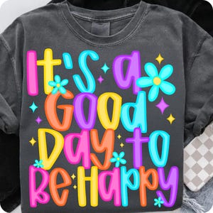 K&ouml;nnte beinhalten: Ein dunkelgraues T-Shirt mit dem Text "It's a good day to be happy" in bunten Buchstaben. Der Text ist von kleinen wei&szlig;en Sternen und bunten Blumen umgeben.