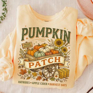 Puede incluir: Una sudadera de manga larga de color amarillo p&aacute;lido con un gr&aacute;fico de un carro de madera lleno de calabazas. El texto dice PUMPKIN PATCH, FRESH PICKED, HAYRIDES, APPLE CIDER y HARVEST DAYS, junto con girasoles, hojas y una paca de heno.