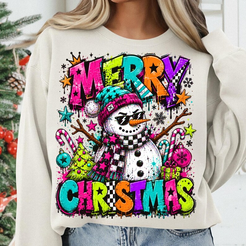 Puede incluir: Una sudadera de cuello redondo color crema con un estampado gr&aacute;fico vibrante de un mu&ntilde;eco de nieve con gafas de sol, un gorro azul cerceta y una bufanda a cuadros. El texto MERRY CHRISTMAS aparece en letras de estilo grafiti multicolor.
