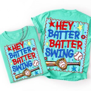 K&ouml;nnte beinhalten: Zwei t&uuml;rkisfarbene T-Shirts mit dem Text "HEY BATTER BATTER SWING" in Rot und Blau. Das Design umfasst einen Baseball, einen Schl&auml;ger und einen Handschuh. Die Shirts haben einen Rundhalsausschnitt und kurze &Auml;rmel. Eine silberne Kette ist auf einem Shirt drapiert.