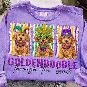 May include: A purple sweatshirt featuring three Golden Doodle dogs in Mardi Gras attire. Each dog wears a different festive accessory: a jester hat, a mask, and a top hat. The text "GOLDENDOODLE Through The beads" is embroidered below the dogs.