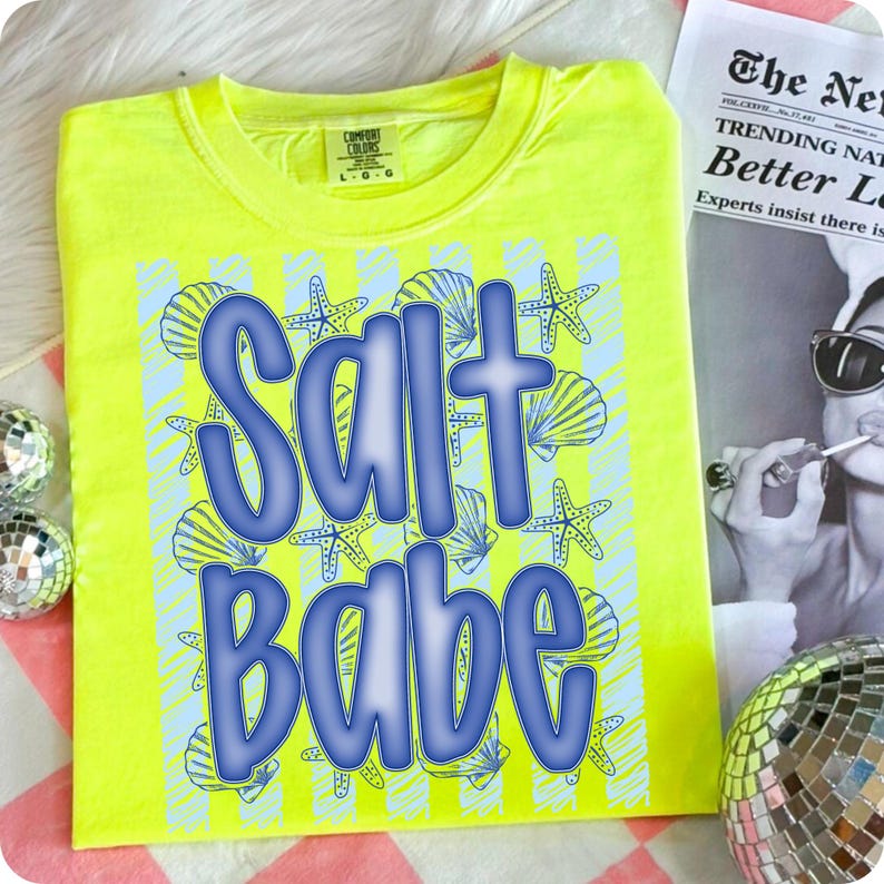 K&ouml;nnte beinhalten: Ein neongelbes T-Shirt mit den Worten "Salt Babe" in Blau, umgeben von blauen Muscheln und Seesternen. Das Shirt ist von der Marke Comfort Colors. Eine Zeitung und eine Discokugel befinden sich im Hintergrund.