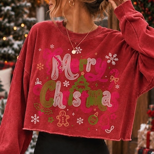 May include: A red long-sleeve cropped sweatshirt featuring the text Merry Christmas in a wavy font. The graphic includes a green Christmas tree, a gingerbread man, a candy cane, holly leaves, and snowflakes in shades of pink and green.