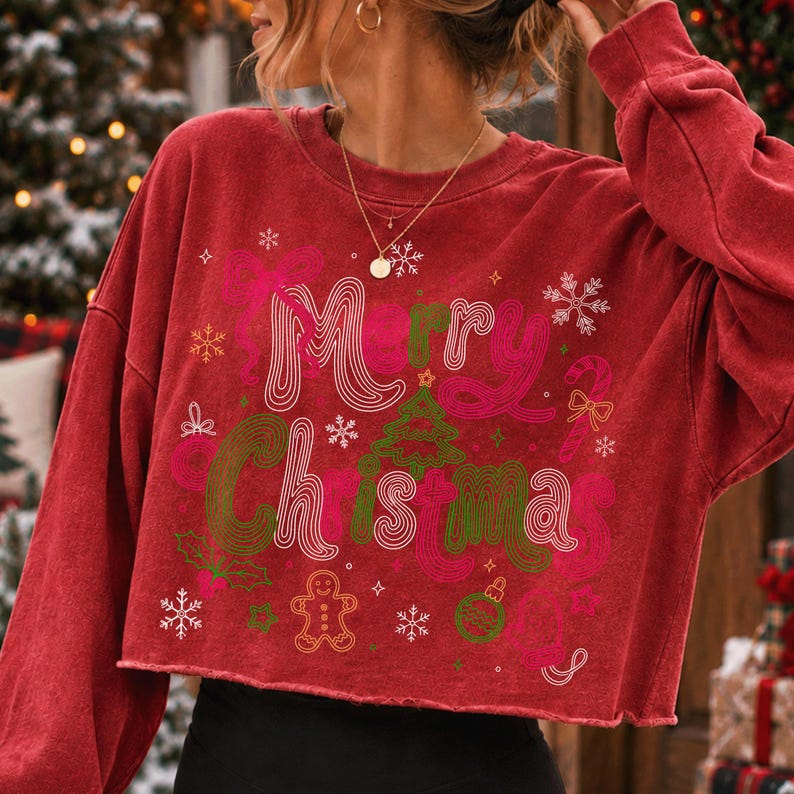 May include: A red long-sleeve cropped sweatshirt featuring the text Merry Christmas in a wavy font. The graphic includes a green Christmas tree, a gingerbread man, a candy cane, holly leaves, and snowflakes in shades of pink and green.