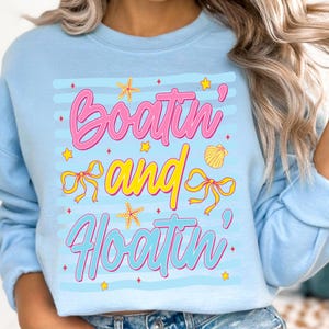 以下が含まれることがあります： ピンクと青の文字で「Boatin' and Floatin'」と書かれた、水色のスウェットシャツ。デザインには、黄色のヒトデ、リボン、貝殻が含まれています。スウェットシャツはクルーネックで長袖です。