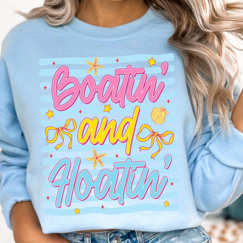 以下が含まれることがあります： ピンクと青の文字で「Boatin' and Floatin'」と書かれた、水色のスウェットシャツ。デザインには、黄色のヒトデ、リボン、貝殻が含まれています。スウェットシャツはクルーネックで長袖です。