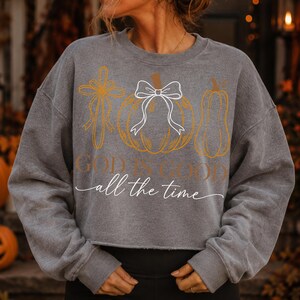 Puede incluir: Una sudadera corta de color gris carb&oacute;n con un estampado gr&aacute;fico de una cruz dorada, una calabaza dorada con un lazo blanco y una calabaza alargada dorada. El texto "GOD IS GOOD" est&aacute; impreso en dorado, con "all the time" en cursiva blanca.