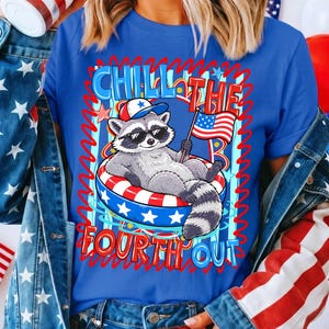 Op de afbeelding: Blauw T-shirt met een cartoon wasbeer met een zonnebril en een patriotische hoed, liggend in een rood, wit en blauw zwemband. Het shirt heeft de tekst "Chill the Fourth Out" in rood en blauw.
