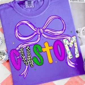 May include: A purple Comfort Colors t-shirt with a large bow design and the word "CUSTOM" in colorful, patterned letters. The bow is purple and white striped. The letters are decorated with polka dots, leopard print, and other patterns.