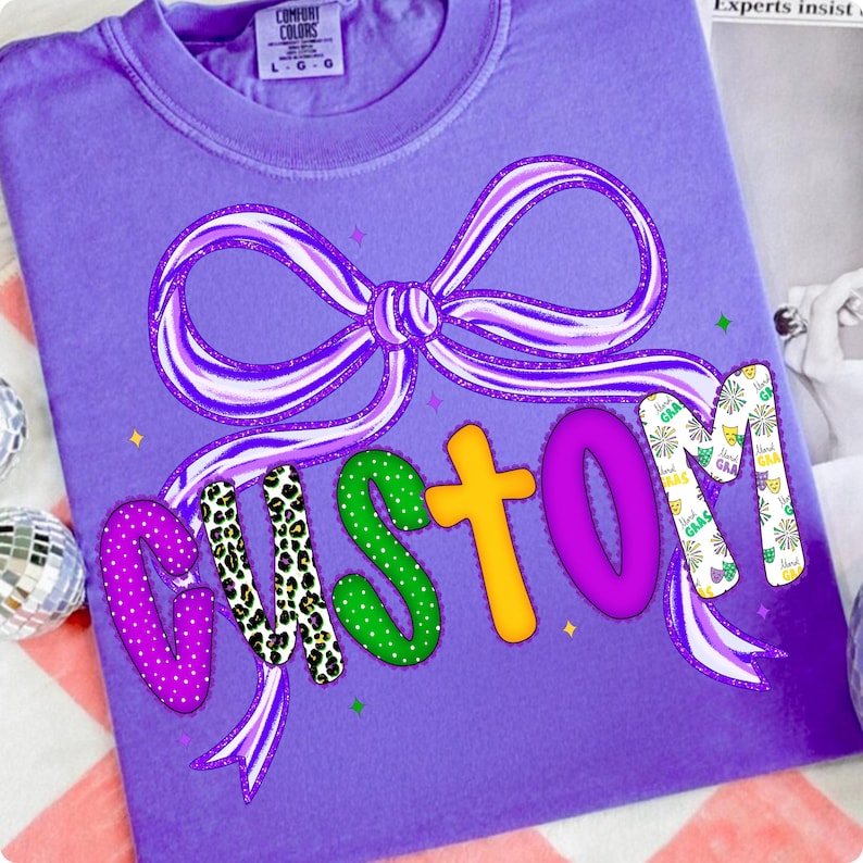 May include: A purple Comfort Colors t-shirt with a large bow design and the word "CUSTOM" in colorful, patterned letters. The bow is purple and white striped. The letters are decorated with polka dots, leopard print, and other patterns.