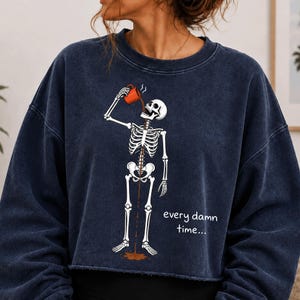 May include: A dark navy blue crewneck sweatshirt featuring a graphic of a white skeleton drinking from an orange mug. The coffee flows through the skeleton's ribcage and out the bottom. The text "every damn time..." is printed in white.