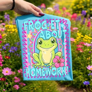K&ouml;nnte beinhalten: Hellblaues T-Shirt mit einem Cartoon-Frosch und dem Text "FROGET ABOUT HOMEWORK!". Der Frosch ist gr&uuml;n mit einer rosa Blume, umgeben von einem rosa und blauen Design. Das T-Shirt wird von einer Person gehalten.