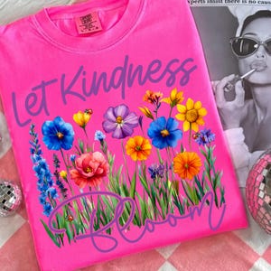May include: A bright pink t-shirt with the words "Let Kindness Bloom" in a cursive font. The shirt features a colorful floral design with blue, yellow, orange, and purple flowers. The shirt is made of Comfort Colors material.