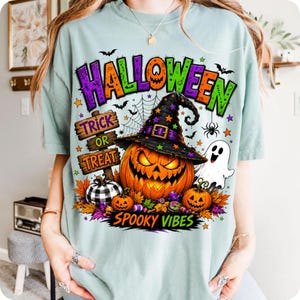 May include: A light green t-shirt featuring a graphic print with the text HALLOWEEN, TRICK OR TREAT, and SPOOKY VIBES. The design includes a large jack-o'-lantern wearing a witch hat, a white ghost, a buffalo plaid pumpkin, and colourful autumn leaves.