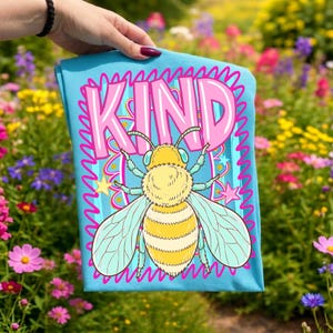 May include: A light blue t-shirt featuring a cartoon bee with yellow and white stripes, light blue wings, and the word "KIND" in pink. The design includes stars and a wavy border. The shirt is held up in front of a colorful flower garden.