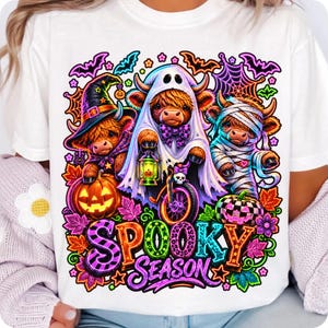 Może przedstawiać: Biały t-shirt z kolorową grafiką przedstawiającą trzy krowy rasy Highland przebrane na Halloween. Jedna ma kapelusz czarownicy, druga prześcieradło ducha z latarnią, a trzecia jest owinięta jak mumia. Wz&oacute;r zawiera dynie, nietoperze oraz napis SPOOKY SEASON.