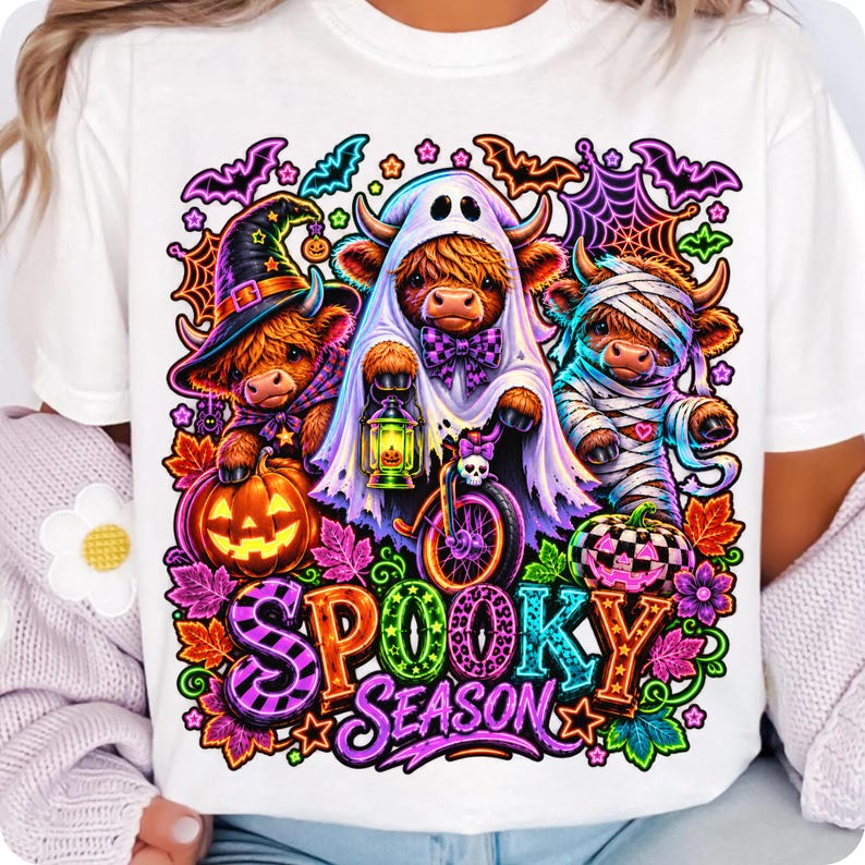 Może przedstawiać: Biały t-shirt z kolorową grafiką przedstawiającą trzy krowy rasy Highland przebrane na Halloween. Jedna ma kapelusz czarownicy, druga prześcieradło ducha z latarnią, a trzecia jest owinięta jak mumia. Wz&oacute;r zawiera dynie, nietoperze oraz napis SPOOKY SEASON.