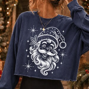 May include: A navy blue long-sleeve cropped sweatshirt featuring a white graphic print of a smiling Santa Claus wearing a hat, accented with holly leaves and starburst shapes. The garment has a crew neckline and a raw, unfinished hemline.