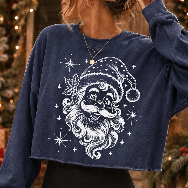 May include: A navy blue long-sleeve cropped sweatshirt featuring a white graphic print of a smiling Santa Claus wearing a hat, accented with holly leaves and starburst shapes. The garment has a crew neckline and a raw, unfinished hemline.