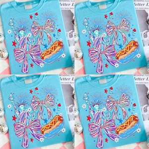 May include: Four light blue t-shirts with a patriotic design. Each shirt features a graphic of the Statue of Liberty, a bow, a hot dog, fireworks, and stars. The text "RENDING NAT Better L erts insist there" is visible.