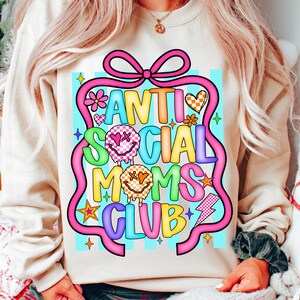 K&ouml;nnte beinhalten: Cremefarbenes Sweatshirt mit einem farbenfrohen Aufdruck "ANTI SOCIAL MOMS CLUB". Das Design zeigt eine rosa Schleife, Blumen, Herzen, Sterne und einen Blitz. Der Text ist in verschiedenen Farben gehalten, darunter Blau, Gelb und Gr&uuml;n.