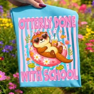 K&ouml;nnte beinhalten: Hellblaues T-Shirt mit einem Cartoon-Otter, der eine herzf&ouml;rmige Sonnenbrille und eine Schleife tr&auml;gt und sich in einem rosa Schwimmring mit einem Getr&auml;nk entspannt. Der Text "OTTERLY DONE WITH SCHOOL" ist rosa.