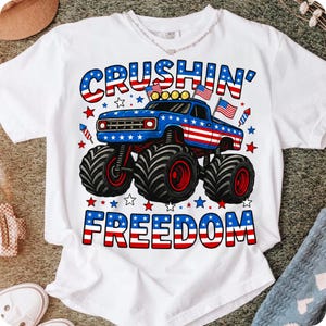 K&ouml;nnte beinhalten: Wei&szlig;es T-Shirt mit einem blauen Monster-Truck-Design mit roten und wei&szlig;en Akzenten, amerikanischen Flaggen und den Worten "Crushin' Freedom" in Rot, Wei&szlig; und Blau. Das Design enth&auml;lt Sterne und Feuerwerk.