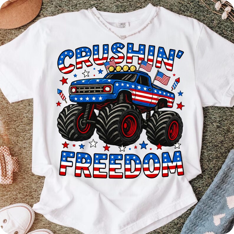 K&ouml;nnte beinhalten: Wei&szlig;es T-Shirt mit einem blauen Monster-Truck-Design mit roten und wei&szlig;en Akzenten, amerikanischen Flaggen und den Worten "Crushin' Freedom" in Rot, Wei&szlig; und Blau. Das Design enth&auml;lt Sterne und Feuerwerk.