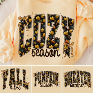 May include: A light peach-colored crewneck sweatshirt featuring the text COZY season in large block letters. The letters are filled with a pattern of yellow, orange, and brown autumn leaves on a black background, accented by two yellow polka-dot bows.