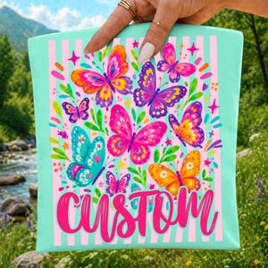 May include: A light mint green tote bag featuring a vertical pink striped pattern and a colourful graphic of various butterflies in shades of pink, purple, orange, and blue, with the word CUSTOM printed in large, bold, bright pink script lettering.