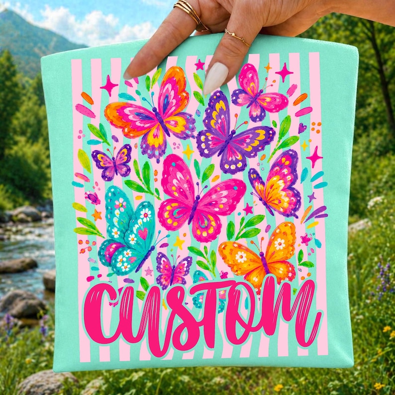 May include: A light mint green tote bag featuring a vertical pink striped pattern and a colourful graphic of various butterflies in shades of pink, purple, orange, and blue, with the word CUSTOM printed in large, bold, bright pink script lettering.