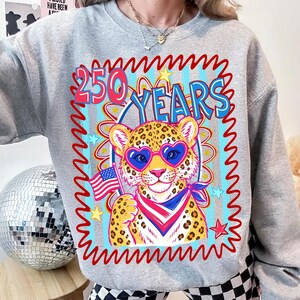 Op de afbeelding: Een grijze sweatshirt met een kleurrijke afbeelding. De afbeelding toont een luipaard met een hartvormige zonnebril en een Amerikaanse vlag. De tekst "250 YEARS" staat in retrostijl boven de luipaard.