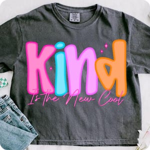 May include: Dark gray t-shirt with the word "Kind" in pink, light blue, and orange bubble letters. The phrase "Is The New Cool" is written below in a cursive font. The shirt is folded with a pair of jeans.