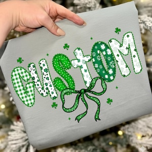 May include: A gray fabric item with the word "CUSTOM" in green and white lettering. The letters have various patterns, including gingham, polka dots, and shamrocks. A green bow and shamrock accents complete the design.