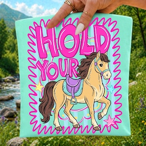 May include: A light teal tote bag with the words "HOLD YOUR" in pink bubble letters. A cartoon horse with a purple saddle is featured below the text, with star accents and a pink zig-zag border.