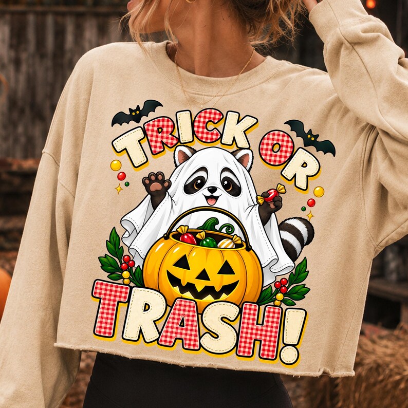 May include: A beige long-sleeve sweatshirt featuring a graphic of a raccoon dressed as a ghost, holding a jack-o'-lantern candy bucket. The text reads "TRICK OR TRASH!" in red and white checkered lettering, surrounded by bats and colourful candy.