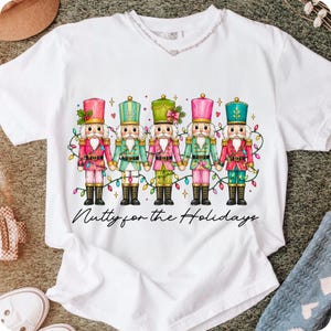 Op de afbeelding: Een wit t-shirt met korte mouwen en een afbeelding van vijf kleurrijke notenkrakers die elkaars hand vasthouden, verbonden door een snoer met veelkleurige kerstverlichting. Onder de figuren staat de tekst Nutty for the Holidays in zwart.