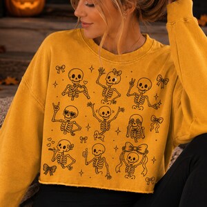 Op de afbeelding: Een mosterdgeel sweatshirt met ronde hals en een zwarte lijntekening van meerdere skeletten in verschillende houdingen, zoals zwaaien, hartjes maken en strikjes dragen, afgewisseld met kleine ster- en striksymbolen op de voorzijde.