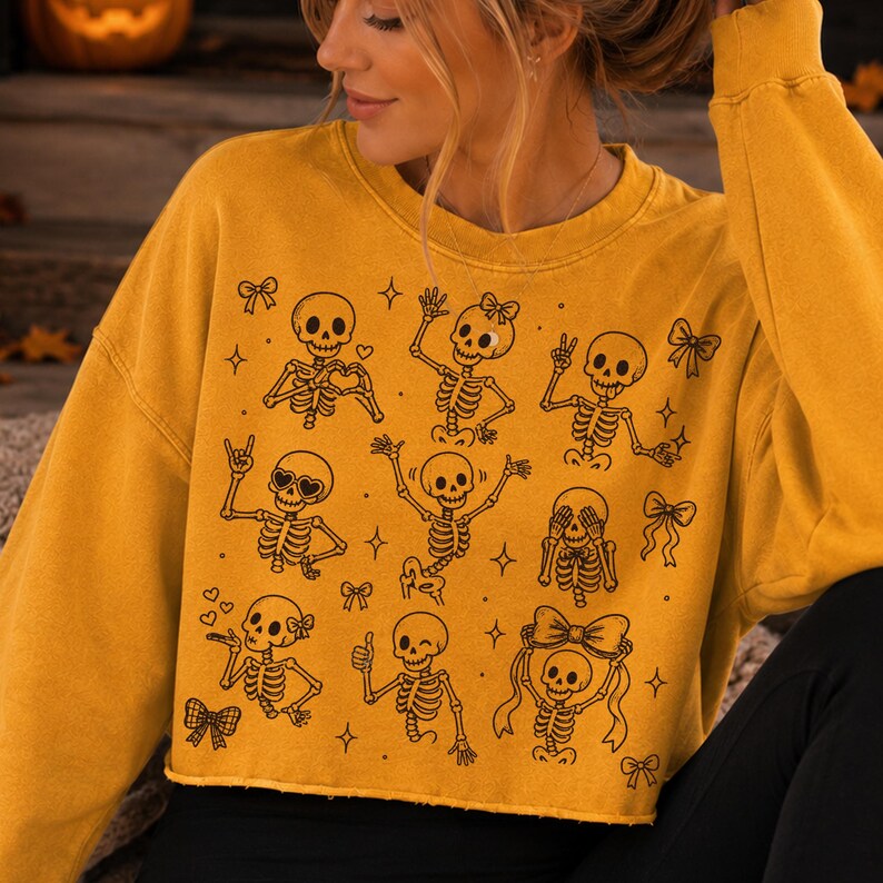 Op de afbeelding: Een mosterdgeel sweatshirt met ronde hals en een zwarte lijntekening van meerdere skeletten in verschillende houdingen, zoals zwaaien, hartjes maken en strikjes dragen, afgewisseld met kleine ster- en striksymbolen op de voorzijde.
