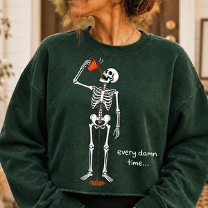 May include: A dark forest green long-sleeve sweatshirt featuring a graphic of a white skeleton drinking from a red mug. The coffee flows through the skeleton's body and spills onto the floor. The text reads every damn time in white letters.